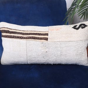 Puede incluir: Cojín decorativo rectangular con diseño patchwork. El cojín presenta un fondo beige claro con una sección de rayas marrones y un detalle bordado en negro. El cojín está colocado sobre una silla azul.