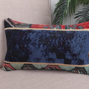 May include: Rectangular decorative pillow with a blue, red, and beige patterned design. The pillow has a dark blue center with a textured appearance, bordered by red and beige sections. The pillow is placed on a beige chair.