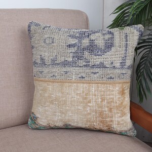 May include: Square decorative pillow with a two-tone design. The top half features a blue and beige pattern, while the bottom half is a textured beige. The pillow is placed on a beige sofa, suggesting a home decor theme.