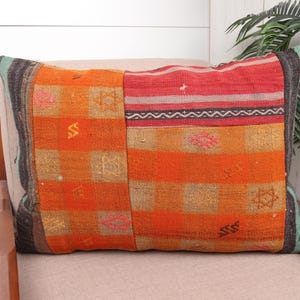 May include: Rectangular decorative pillow with a patchwork design. The pillow features a vibrant mix of orange, red, and tan hues, with geometric and striped patterns. The pillow is placed on a neutral-toned chair.