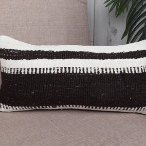 May include: Rectangular pillow with a woven design in black and white. The pillow features horizontal stripes, with a wide black band at the bottom and a white band at the top. The pillow is placed on a neutral-toned surface.