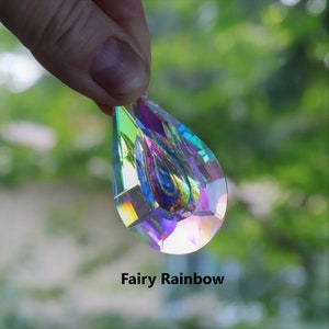 May include: A teardrop-shaped crystal prism with iridescent rainbow colors. The prism is held in a hand against a blurred green background. "Fairy Rainbow" is written below the prism.
