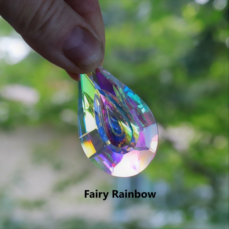 May include: A teardrop-shaped crystal prism with iridescent rainbow colors. The prism is held in a hand against a blurred green background. "Fairy Rainbow" is written below the prism.