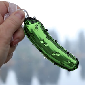 May include: A green glass pickle ornament with small air bubbles throughout. The ornament is shaped like a pickle and has a loop at the top for hanging.