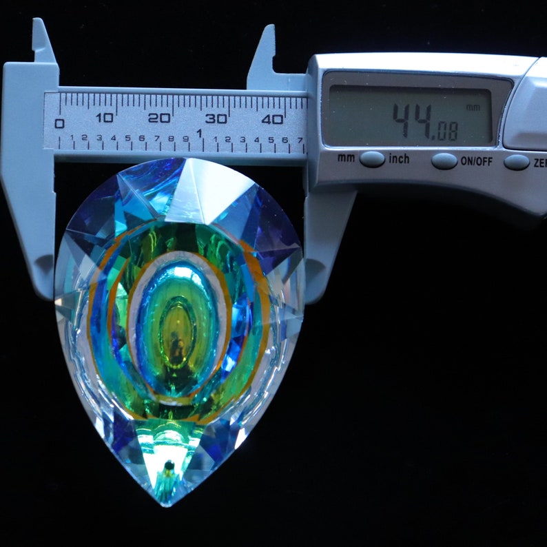 May include: A clear crystal prism with a teardrop shape and a multi-colored iridescent design. The prism is being measured with a caliper, which reads 44.08 mm.