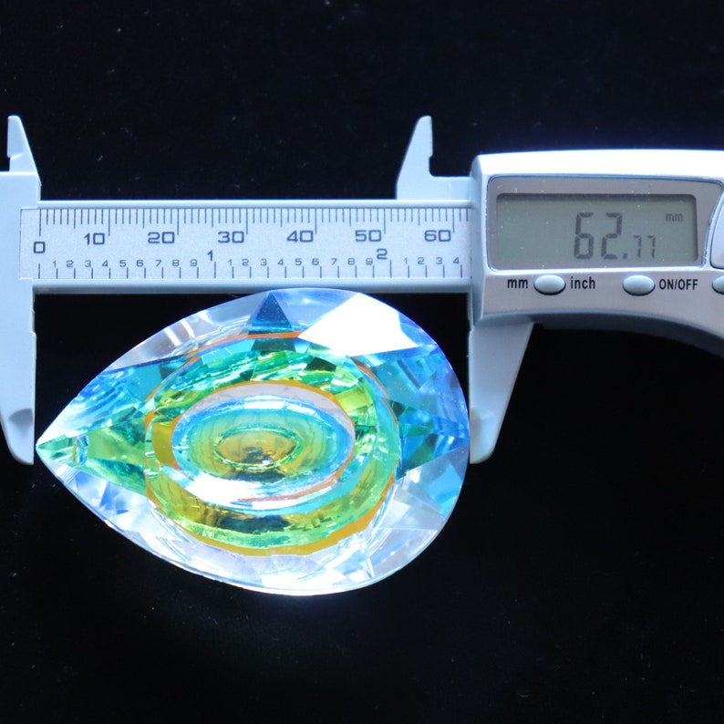 May include: A teardrop-shaped crystal prism with a swirling pattern of green, blue, and yellow. The prism is being measured with a caliper, which reads 62.11 mm.