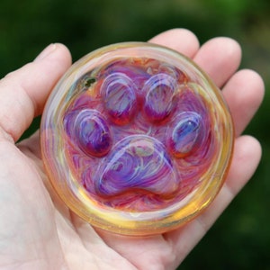 May include: A round glass paperweight with a paw print design. The paw print is made of purple, pink, and white glass, and the background is a swirl of yellow and orange glass.