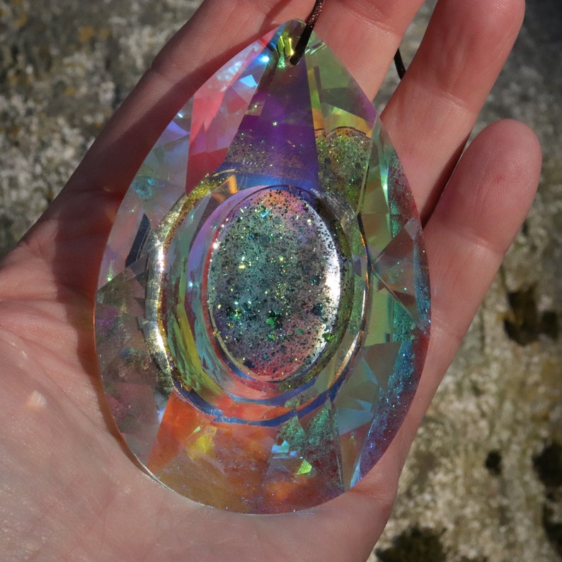 May include: A teardrop-shaped iridescent crystal pendant with a smaller, round, green and silver glitter-filled crystal inside. The pendant is hanging from a brown cord.