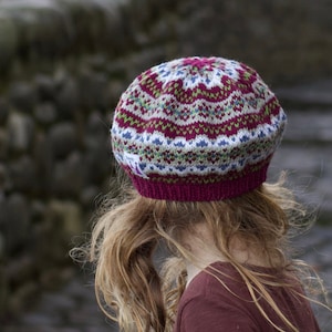 Könnte beinhalten: Eine junge Person trägt eine rote, weiße und grüne gestrickte Mütze mit einem burgunderfarbenen Kleid. Die Person steht auf einem Kopfsteinpflasterweg.