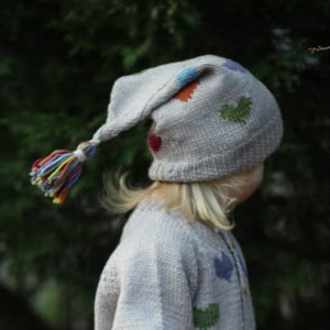 May include: A young person wearing a grey knitted hat with a long, colourful tassel, a grey cardigan with colourful heart designs, and pink wellies. The person is standing in a woodland setting.