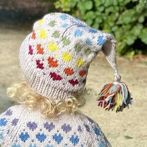 May include: A child wearing a beige knitted jumper with a rainbow heart pattern and a matching hat with a long pointy tip and a colourful pom-pom.