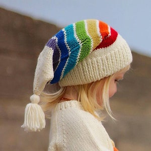Puede incluir: Un niño lleva una gorra blanca de punto con un borde a rayas de arcoíris y un borla blanca. La gorra tiene una solapa de oreja larga y puntiaguda.