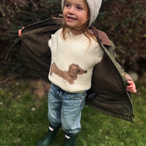 May include: A young child wearing a white knit sweater with a brown dachshund design, a white knit hat, blue jeans, and green rain boots. The child is standing in a grassy area.