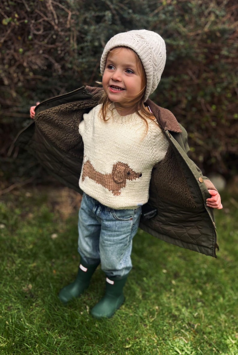 May include: A young child wearing a white knit sweater with a brown dachshund design, a white knit hat, blue jeans, and green rain boots. The child is standing in a grassy area.
