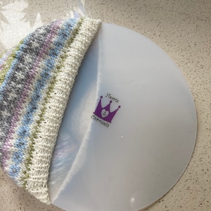 May include: A white round disc with a purple crown and the text "Hearts & Coronets" printed on it. A knitted hat with a light blue, green, and gray pattern is partially covering the disc.
