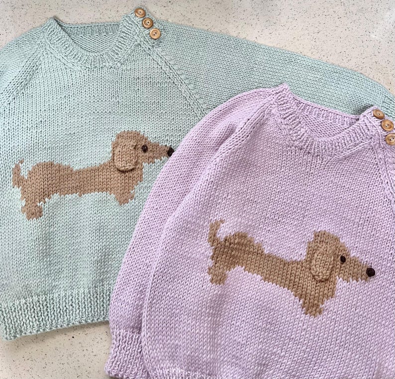 May include: Two knitted sweaters with dachshund designs. One sweater is light green with a brown dachshund, the other is light purple with a brown dachshund.