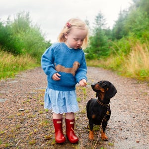 May include: A young girl in a blue sweater with a brown dachshund design is feeding a black dachshund a treat. She is wearing red rain boots and a white skirt. The dog is sitting on a gravel path.