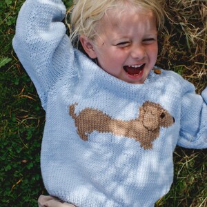 May include: A young child wearing a light blue knitted sweater with a brown dachshund design. The child is lying on their back in a grassy field and laughing.
