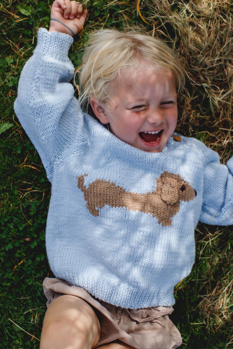 May include: A young child wearing a light blue knitted sweater with a brown dachshund design. The child is lying on their back in a grassy field and laughing.