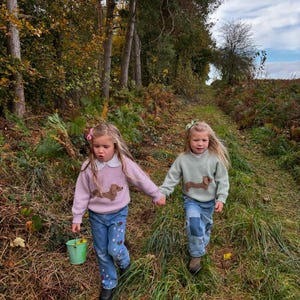 May include: Two young girls wearing sweaters with dachshund dog designs walk along a path in a forest. The girl on the left is wearing a pink sweater and the girl on the right is wearing a green sweater. Both girls are wearing blue jeans.