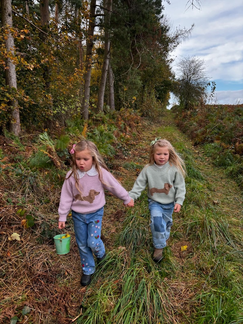 May include: Two young girls wearing sweaters with dachshund dog designs walk along a path in a forest. The girl on the left is wearing a pink sweater and the girl on the right is wearing a green sweater. Both girls are wearing blue jeans.