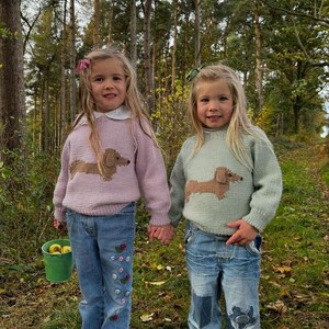 May include: Two young girls wearing knitted sweaters with dachshund designs. The sweaters are in pink and light green. The girls are wearing blue jeans, one with embroidered flowers and the other with patches. They are standing in a forest setting.