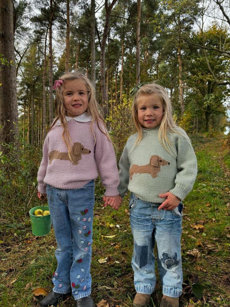 May include: Two young girls wearing knitted sweaters with dachshund designs. The sweaters are in pink and light green. The girls are wearing blue jeans, one with embroidered flowers and the other with patches. They are standing in a forest setting.