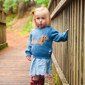 May include: A young girl wearing a blue knitted sweater with a brown dachshund design, a light blue skirt, and red rain boots. She is standing on a wooden bridge with a wooden railing.