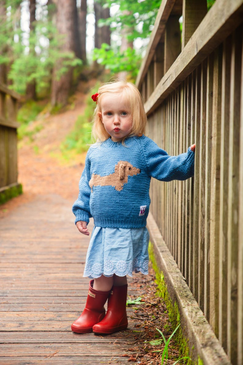 May include: A young girl wearing a blue knitted sweater with a brown dachshund design, a light blue skirt, and red rain boots. She is standing on a wooden bridge with a wooden railing.