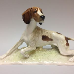 Puede incluir: Una figurita de porcelana de un perro de caza marrón y blanco acostado sobre una base verde. El perro tiene un hocico largo y orejas caídas.