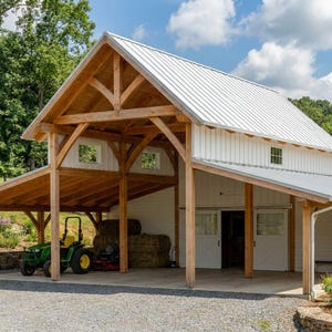 May include: A large timber frame barn with white vertical siding and a silver metal roof. The structure features an open-sided bay with exposed wooden beams, housing a green tractor and hay bales. White double doors are visible on the main building.