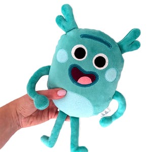 May include: A teal plush toy with a smiling face, large black eyes, and a pink mouth. The toy has two small antennae and two light blue circles on its face. It is being held by a person's hand.