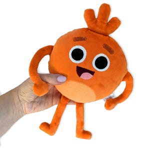 Könnte beinhalten: Oranges Plüschtier mit einem lächelnden Gesicht, schwarzen Augen und einem rosa Mund. Das Spielzeug hat Arme, Beine und eine dreizackige Oberseite. Das Spielzeug wird in einer Hand vor weißem Hintergrund gehalten.
