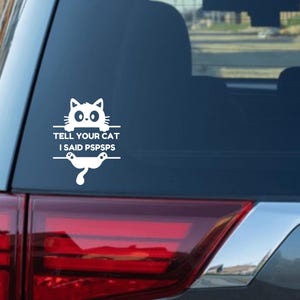 Puede incluir: Adhesivo para coche de vinilo blanco con un gato de dibujos animados y el texto "TELL YOUR CAT I SAID PSPSPS". El adhesivo está diseñado para aplicarse en la ventana de un coche o superficie similar. El gato tiene ojos grandes y una pose juguetona.