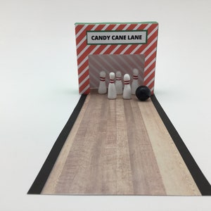 Könnte beinhalten: Ein Miniatur-Bowling-Set mit einer rot-weiß gestreiften Bahn mit der Aufschrift "Candy Cane Lane". Fünf weiße Bowling-Pins sind auf einer Holzbahn aufgestellt, mit einer schwarzen Bowlingkugel am Ende.