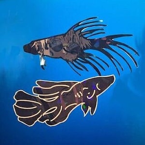 May include: Two decorative fish cutouts. The top fish is dark with spiky fins and a textured pattern. The bottom fish is a similar design, with a curved body and a smiling face. Both are set against a bright blue background.