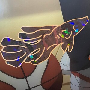 May include: A holographic sticker featuring a stylized hand gesture. The sticker has a copper outline and iridescent accents that reflect multiple colors. The background shows a basketball and other elements.