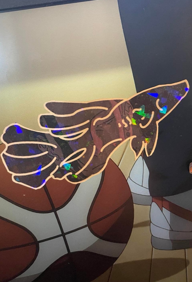 May include: A holographic sticker featuring a stylized hand gesture. The sticker has a copper outline and iridescent accents that reflect multiple colors. The background shows a basketball and other elements.