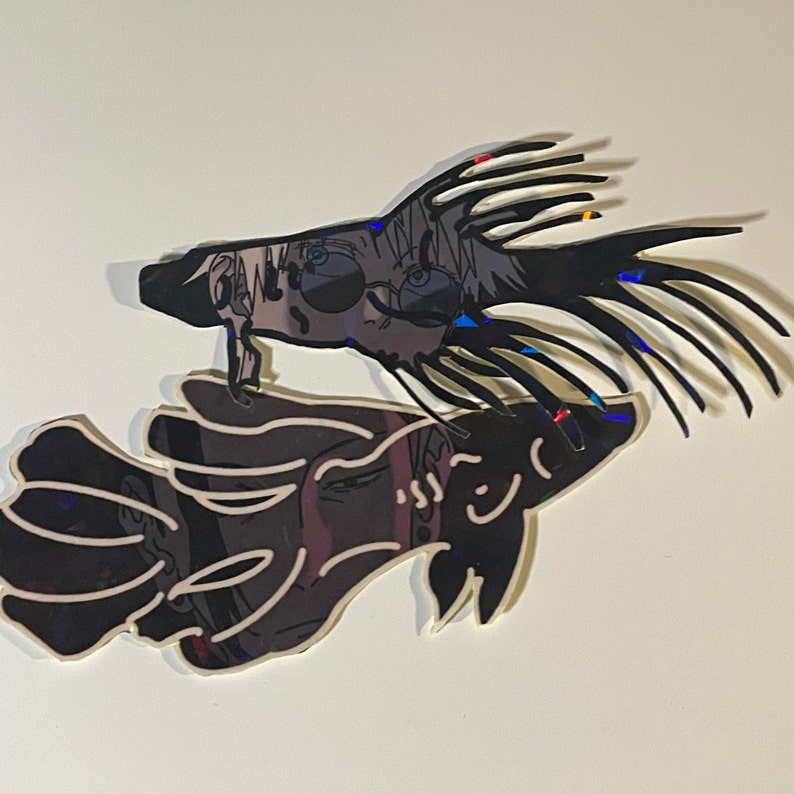 May include: Two mirrored, stylized fish-shaped art pieces. The top fish is black with a detailed illustration of a person wearing sunglasses. The bottom fish is a mirrored image of the top fish, with a similar illustration. The art pieces are mounted on a white background.