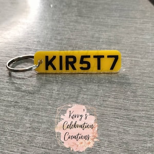 May include: A yellow acrylic keyring with the registration number "KIR5T7" in black. The keyring is attached to a silver split ring. The background is a grey, textured surface. The keyring is a personalized item.