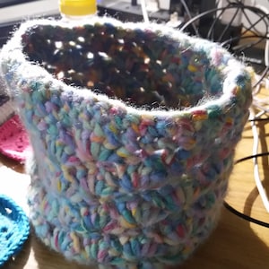 Op de afbeelding: Een gehaakte mand met een kleurrijk, veelkleurig garenontwerp. De mand is gemaakt van zacht, pluizig garen en heeft een lichtblauwe basis.