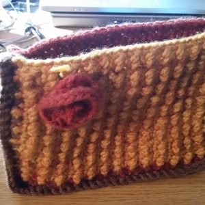 May include: A crocheted pouch with a brown, orange, and red color scheme. The pouch has a small red crocheted flower on the front.