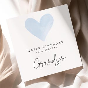 May include: A white birthday card with a light blue watercolor heart. The card reads "Happy Birthday to a Special Grandson" in black text. The card is on a cream-colored fabric background.