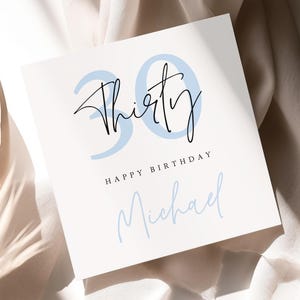 May include: A square white greeting card featuring the large light blue number 30 in the background with the word Thirty written in black cursive script over it. Below this, the text HAPPY BIRTHDAY is printed in black, followed by the name Michael.