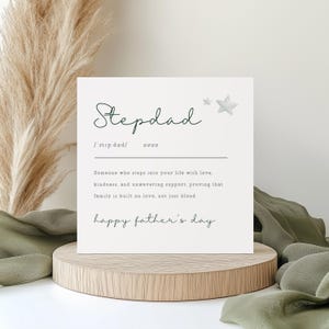 May include: An off-white card with the word "Stepdad" in script, with a definition and the phrase "happy father's day". The card is displayed on a wooden stand, with decorative elements in the background.