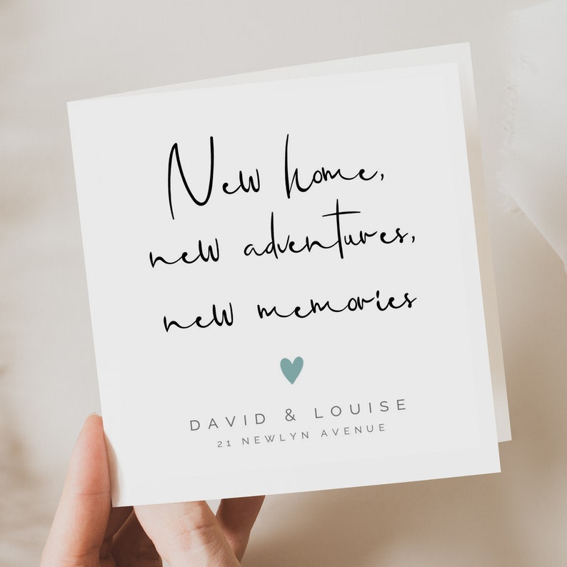May include: A white greeting card with the text "New home, new adventures, new memories" in black script. Below the text is a small teal heart. The card also includes the names "David & Louise" and the address "21 Newlyn Avenue".