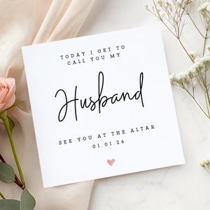 May include: A white square card with the words "TODAY I GET TO CALL YOU MY Husband" in black script. Below, it reads "SEE YOU AT THE ALTAR 01.01.26" with a small pink heart. The card is decorated with flowers.