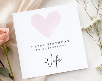 May include: A white greeting card with a light pink watercolor heart. The card reads "HAPPY BIRTHDAY TO MY BEAUTIFUL Wife" in black text. A pink rose and white flowers are in the background.