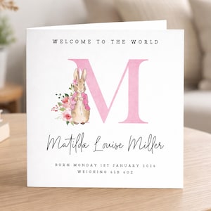 May include: A square white greeting card featuring the text WELCOME TO THE WORLD, a large pink letter M, and a watercolor illustration of a rabbit in a pink jacket with flowers. Below is the name Matilda Louise Miller, birth date, and 6lb 6oz.