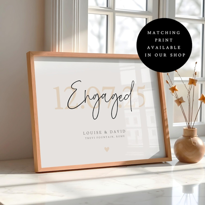 Puede incluir: Una impresi&oacute;n enmarcada con la palabra "Engaged" en escritura elegante, junto con la fecha "12.07.25" y los nombres "Louise & David". La impresi&oacute;n est&aacute; en un marco de madera clara, con un peque&ntilde;o s&iacute;mbolo de coraz&oacute;n en la parte inferior. Un c&iacute;rculo negro con texto est&aacute; en la esquina superior derecha.